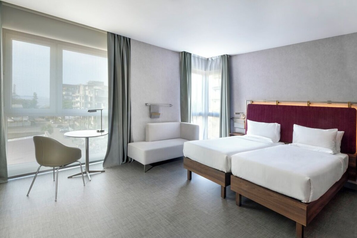 A hotel room with two beds, a small table, a chair, and a view of the city.