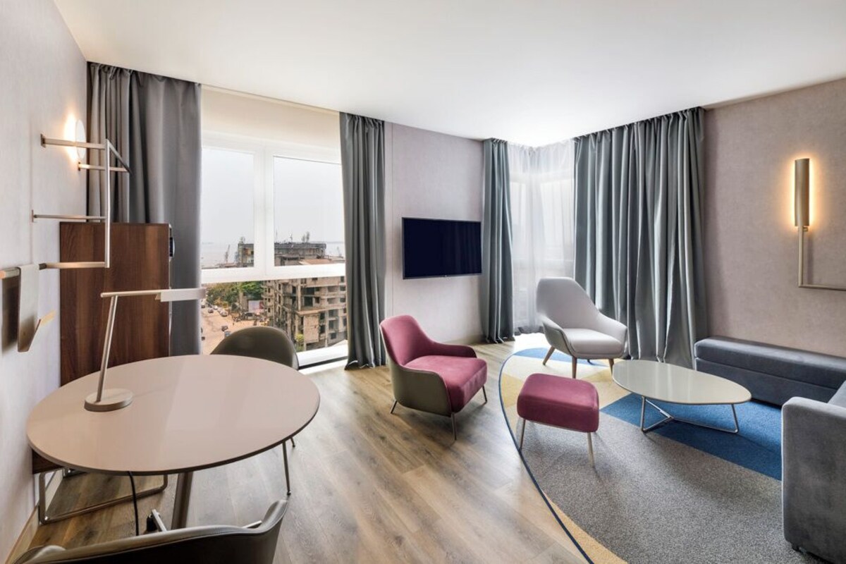 A modern hotel room with a round table, two armchairs, a sofa, a flat-screen TV, and a city view.