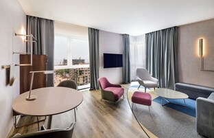 Una habitación de hotel moderna con una mesa redonda, dos sillones, un sofá, un televisor de pantalla plana y vista a la ciudad.