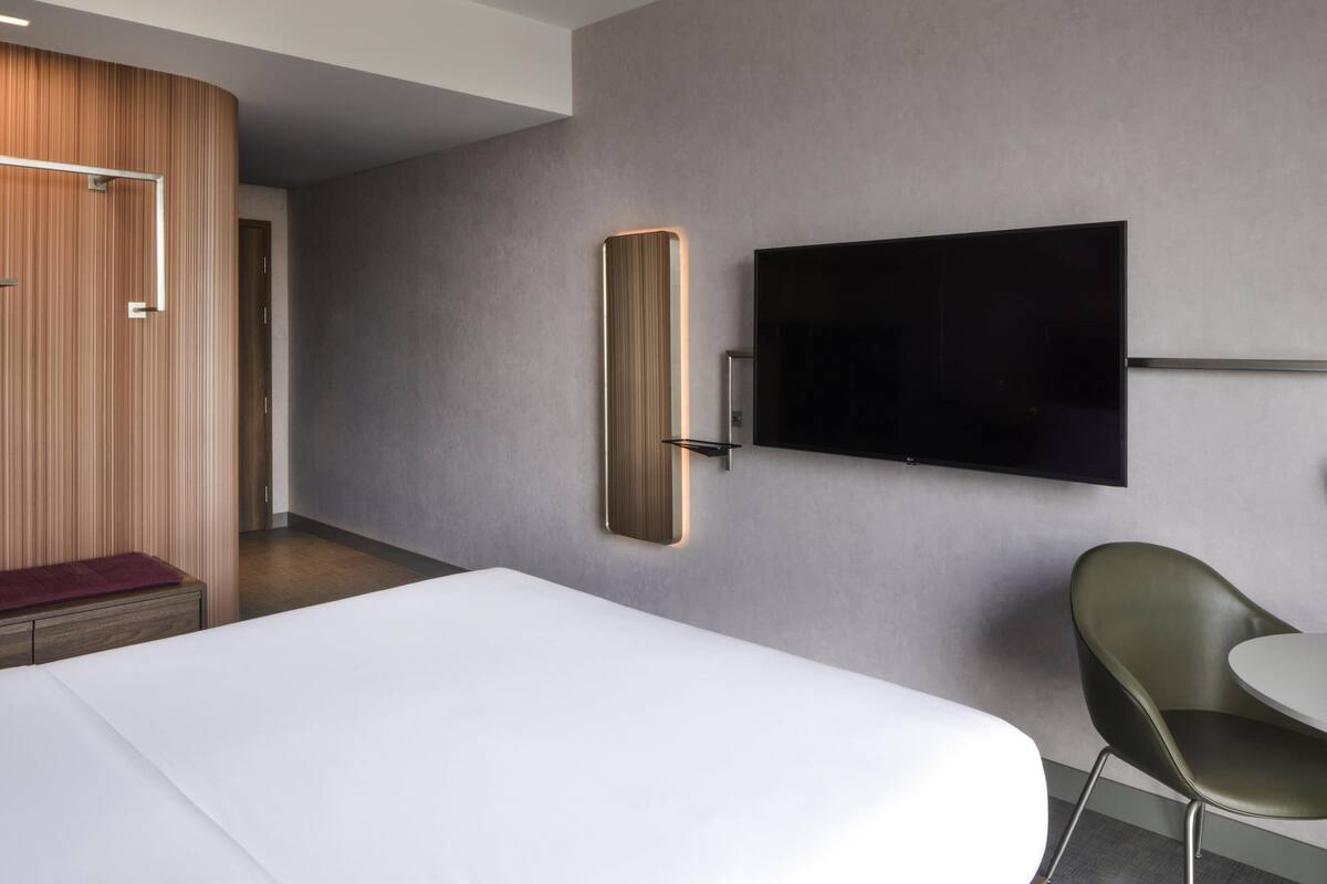 A modern hotel room with a large bed, a flat-screen TV mounted on the wall, a wooden wardrobe, and a green chair.