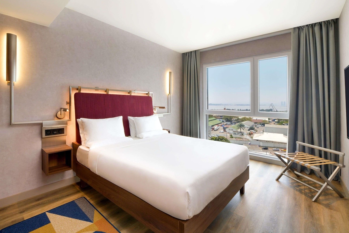 A hotel room with a large bed, a nightstand, a mirror, a wall-mounted light, and a view of a cityscape through large windows.