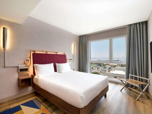 A hotel room with a large bed, a nightstand, a mirror, a wall-mounted light, and a view of a cityscape through large windows. - Novotel Kinshasa La Gombe (Kinshasa)