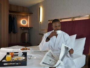 A hotel room with a bed, a tray with breakfast items, a newspaper, and a robe-wearing person. - Novotel Kinshasa La Gombe (Kinshasa)