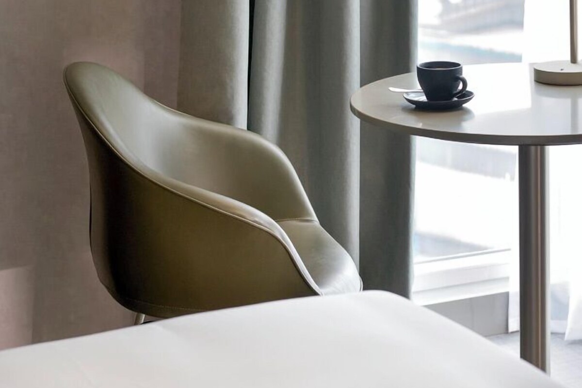 A modern hotel room with a curved chair, a small round table with a cup, and a view of a building through large curtains.