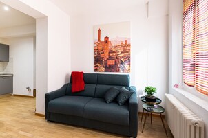 A modern living room with a dark grey sofa, a red throw blanket, a cityscape painting, a small round table with a potted plant, and a radiator.