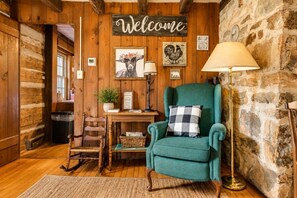 A rustic interior with a wooden wall, a welcome sign, a green armchair, a lamp, and a table with a plant. - Riverfront Cabin! Mini Highland Cows & Kayaks (DB) (Luray)