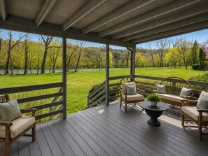 A covered outdoor deck with wooden furniture and a view of a lake and trees.