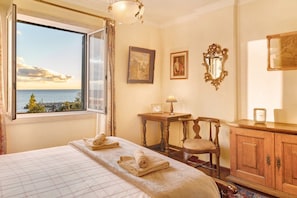 A hotel room with a bed, a wooden chair, a small table with a lamp, a clock, and a view of the sea.