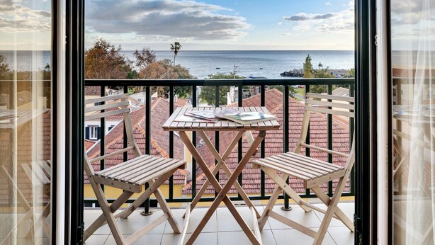 Un balcón con dos sillas de madera, una mesita y vista al mar y edificios.