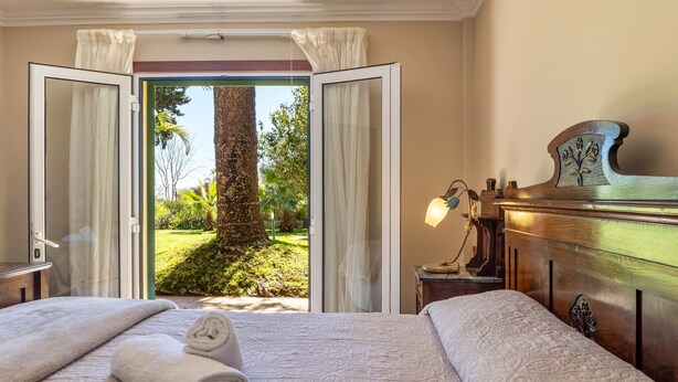 Camera d'albergo con un letto, un comodino, una lampada e vista su un giardino attraverso una porta aperta.