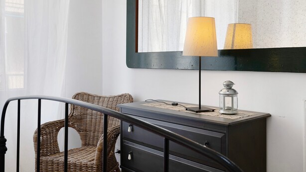 A room with a wicker chair, a dresser with a lamp, and a mirror on the wall.