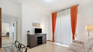 A living room with a sofa, a TV on a cabinet, a dining table with chairs, and a window with curtains.