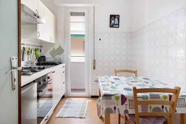 Een kleine keuken met witte kastjes, een kookplaat en een eettafel met een tafelkleed met een geometrisch keukenmotief.