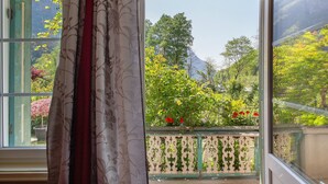 A room with a large window, a view of a garden with trees and flowers, and a curtain partially drawn. - Apartment 'Villa Stanga' with Mountain View, Private Garden and Wi-Fi (Sedico)