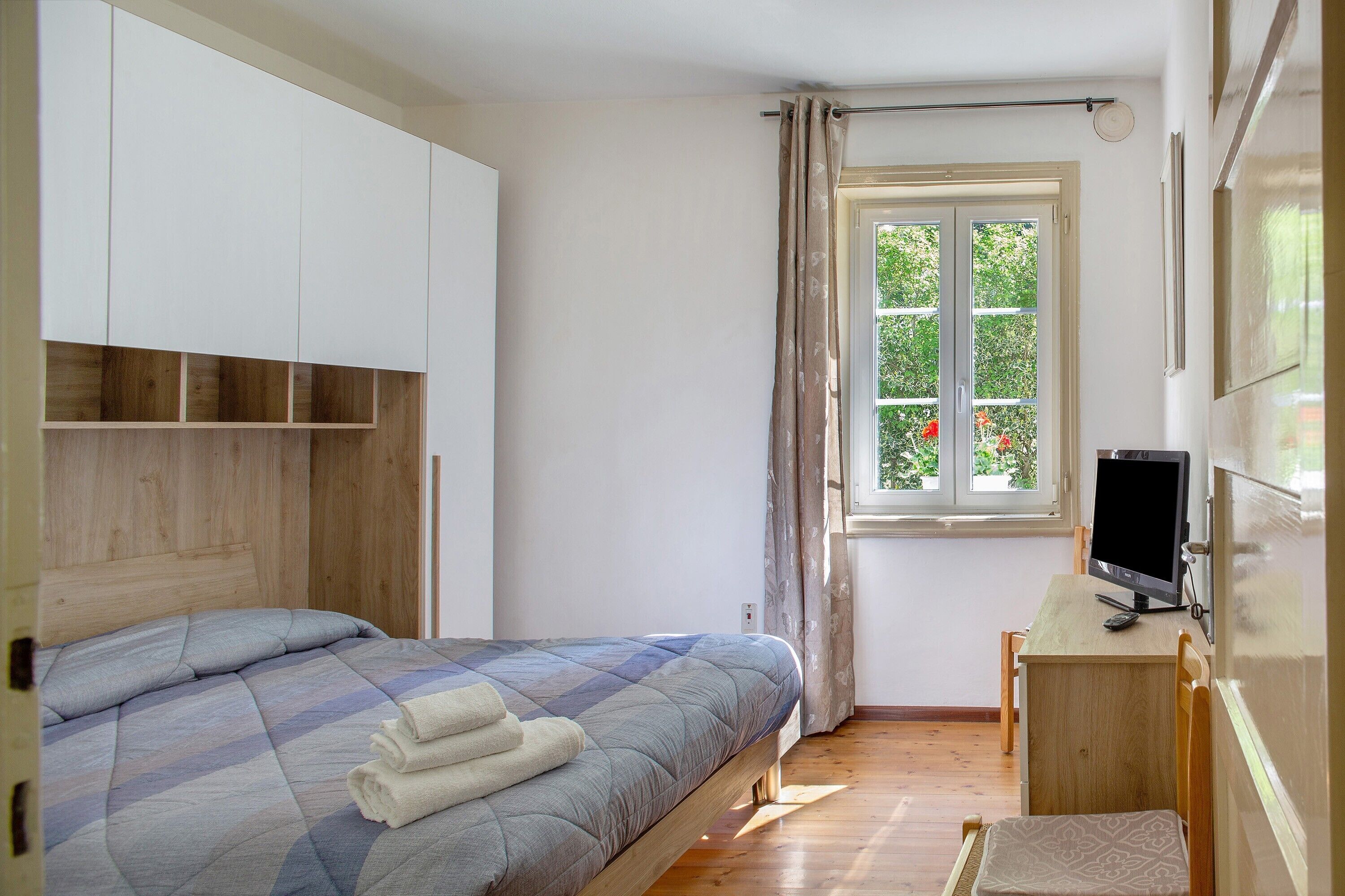 A bedroom with a bed, a desk with a computer, a window with curtains, and a wardrobe.
