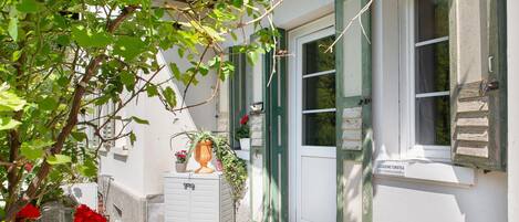 A white building with green shutters, a door, and a window. There is a sign on the wall and a potted plant on the ground.