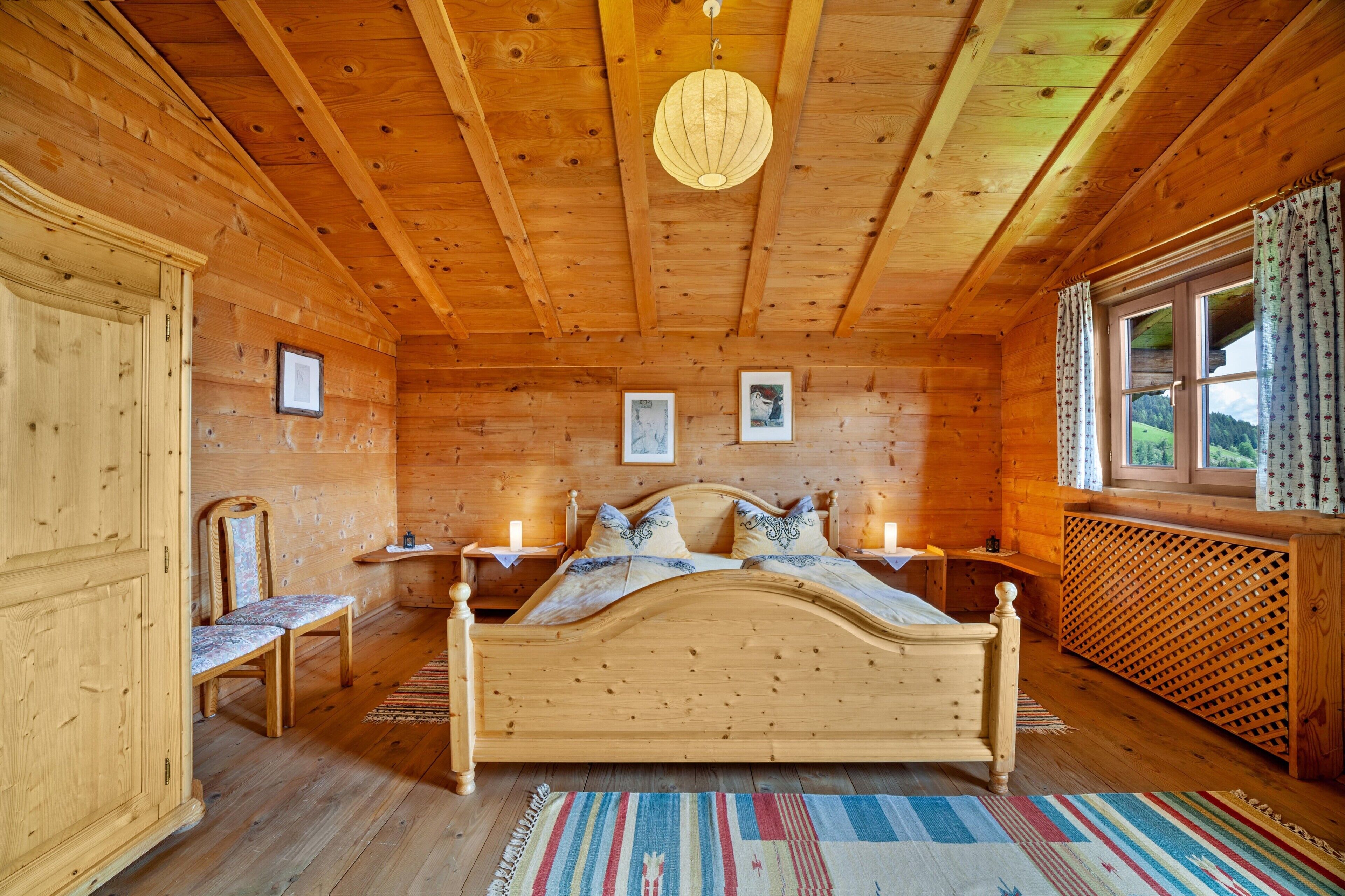 Ein Schlafzimmer einer Holzhütte mit einem Bett, Nachttischen, einem Sessel und einem Fenster mit Vorhängen.