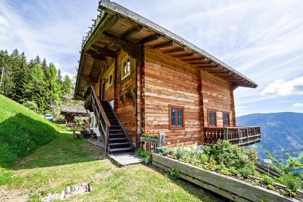 Eine Holzhütte mit einer Treppe zum Eingang, umgeben von einem Garten und Blick auf die Berge.