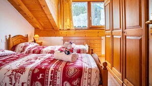 Camera da letto in una baita di legno con due letti, una finestra con tende, un armadio in legno e un comodino con una lampada.