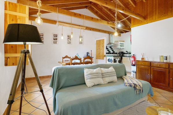 A room with a bed, wooden beams, a tripod lamp, and a kitchen in the background.