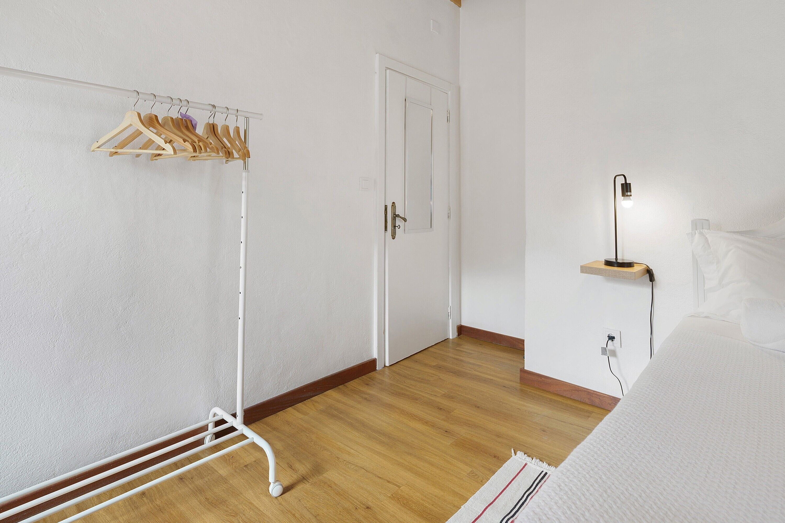Una habitación con suelo de madera, una puerta blanca, un perchero con ganchos, una mesita de noche con lámpara y una cama con ropa de cama blanca.