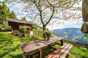 A wooden picnic table with benches, a rustic wooden cabin, and a mountainous landscape in the background.