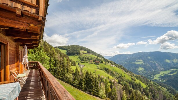 Un balcon en bois, agrémenté d’une table et de chaises, qui offre une vue sur un paysage montagneux, avec des champs verdoyants et des maisons dispersées.