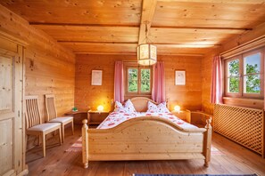 A wooden cabin bedroom with a bed, two chairs, a nightstand with a lamp, and a window with curtains.