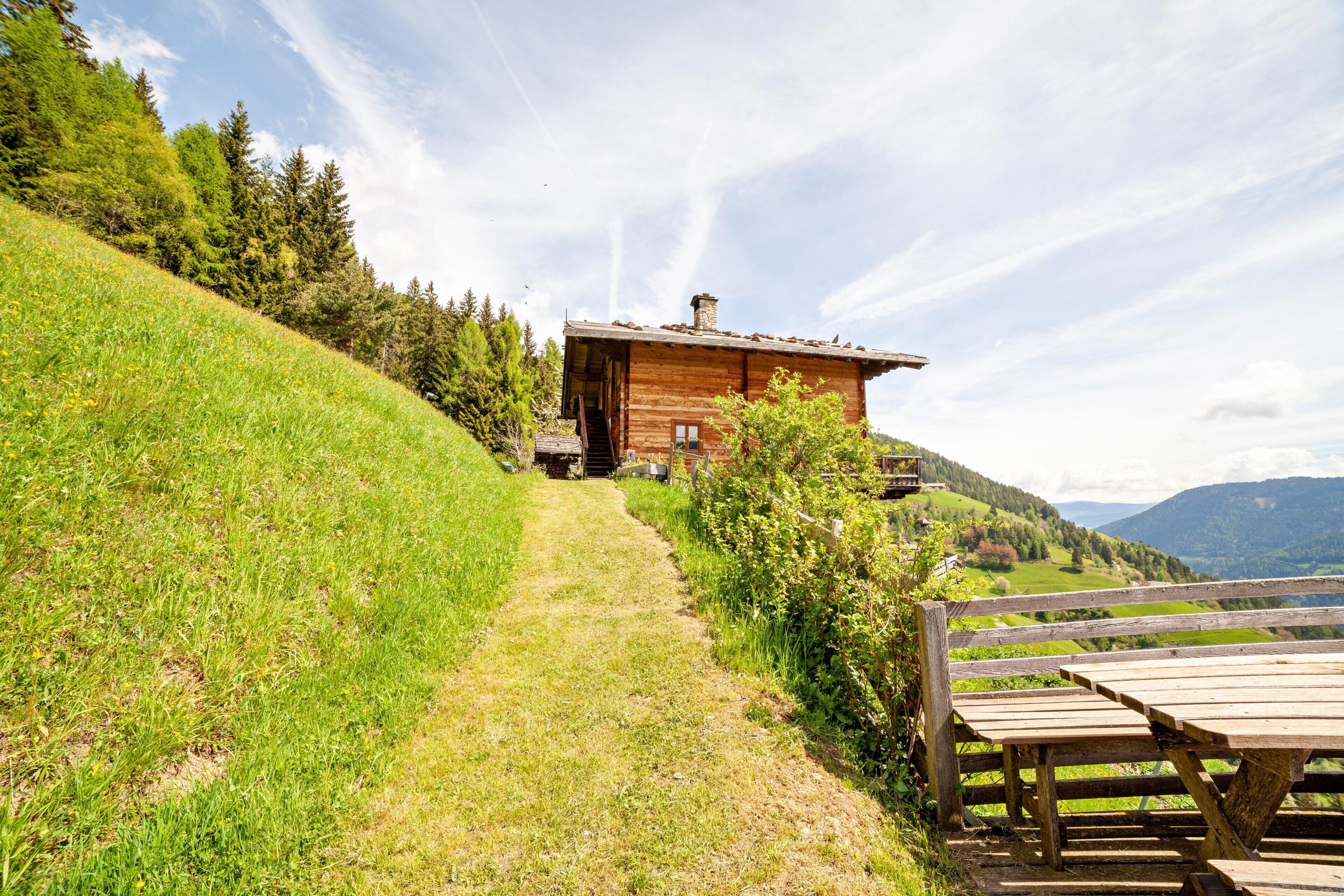 A wooden cabin with a dirt path leading to it, surrounded by greenery and hills.
