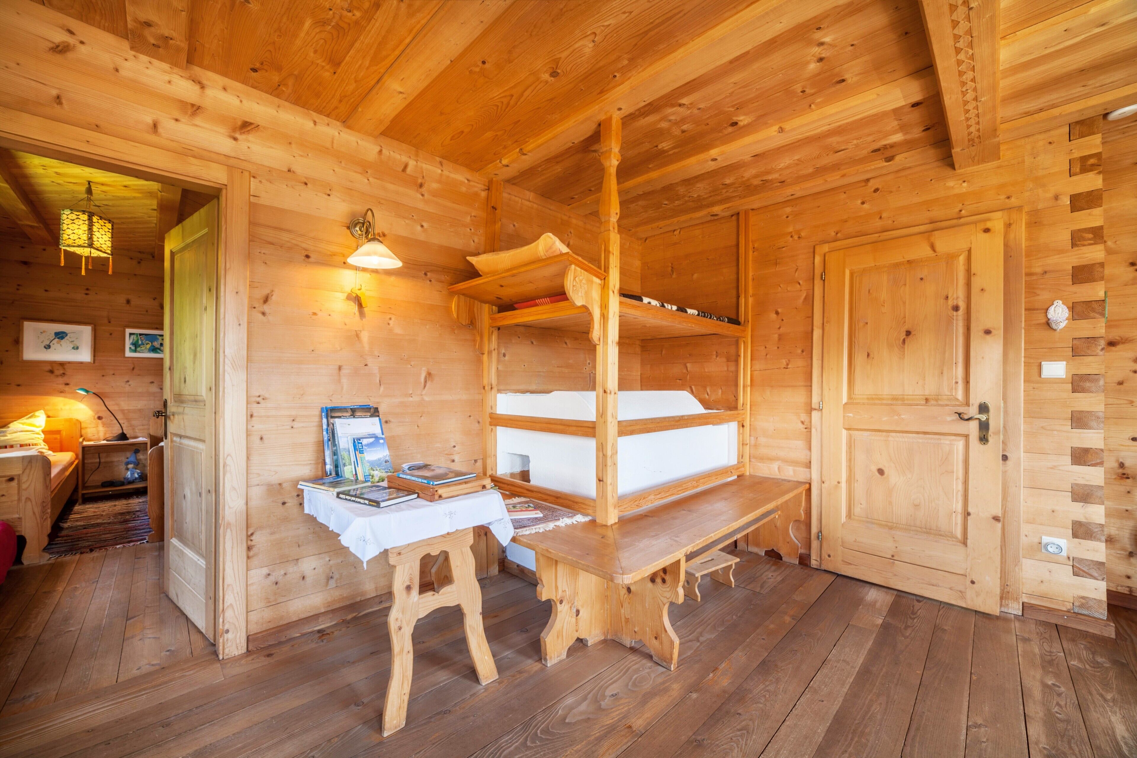 A wooden cabin interior with a bunk bed, a desk with a chair, and a door.
