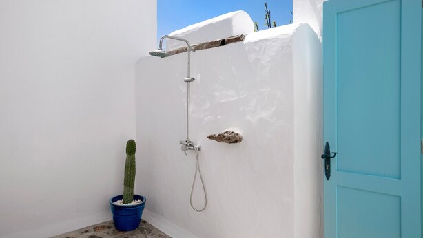 Een smalle buitenruimte met een witte muur, een blauwe deur, een douchekop, een cactus in een blauwe pot en een stenen vloer.