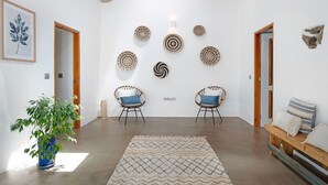 Een kamer met een grote potplant, twee rieten stoelen met blauwe kussens, een houten bank met gestreepte kussens en een kamerkleed met dessin.