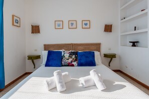 Een opgemaakt bed met witte lakens en blauwe kussens, een klein nachtkastje met een groene lamp, en een muur met ingelijste foto's.