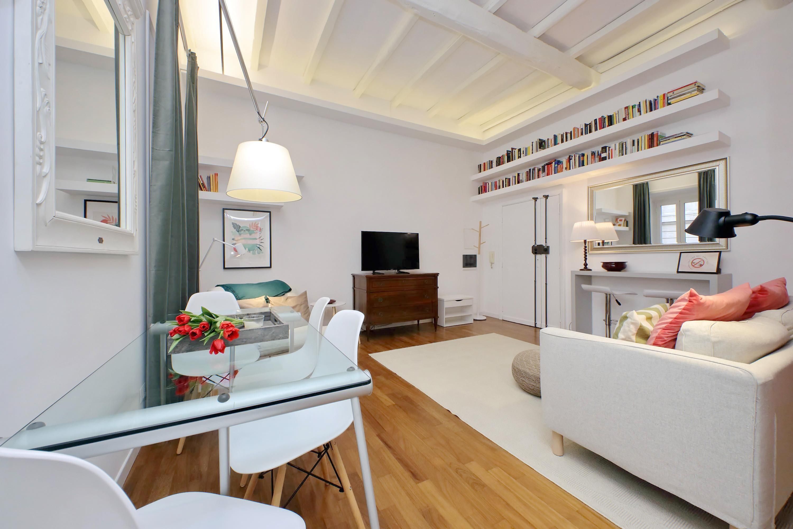 A modern living room with a glass dining table, white sofas, a TV, bookshelves, and a mirror.