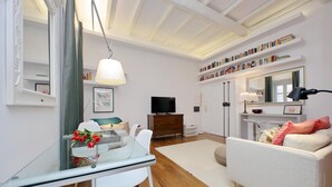 A modern living room with a glass dining table, white sofas, a TV, bookshelves, and a mirror. - 4bnb - Serpenti Delightful Apartment (Rome)