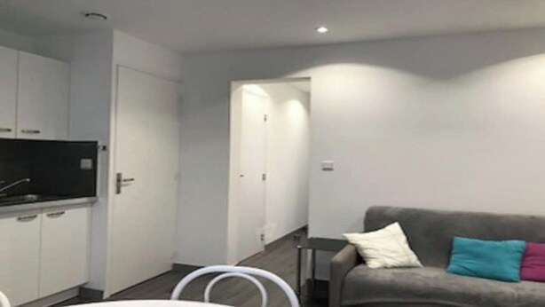 A compact living space with a kitchenette, a grey sofa with colorful cushions, and a hallway leading to a door. - Pacific Apartment (Fort-Mahon-Plage)