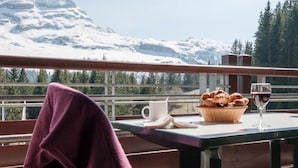 Un balcon avec une table préparée pour le petit-déjeuner, comprenant du pain, un verre de vin rouge et une tasse. Une serviette violette est accrochée à la balustrade.