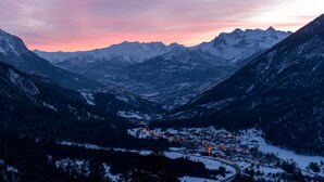 A snow-covered mountain town at dusk with illuminated buildings and a winding road. - Village Club du Soleil de Montgenèvre (Montgenevre)