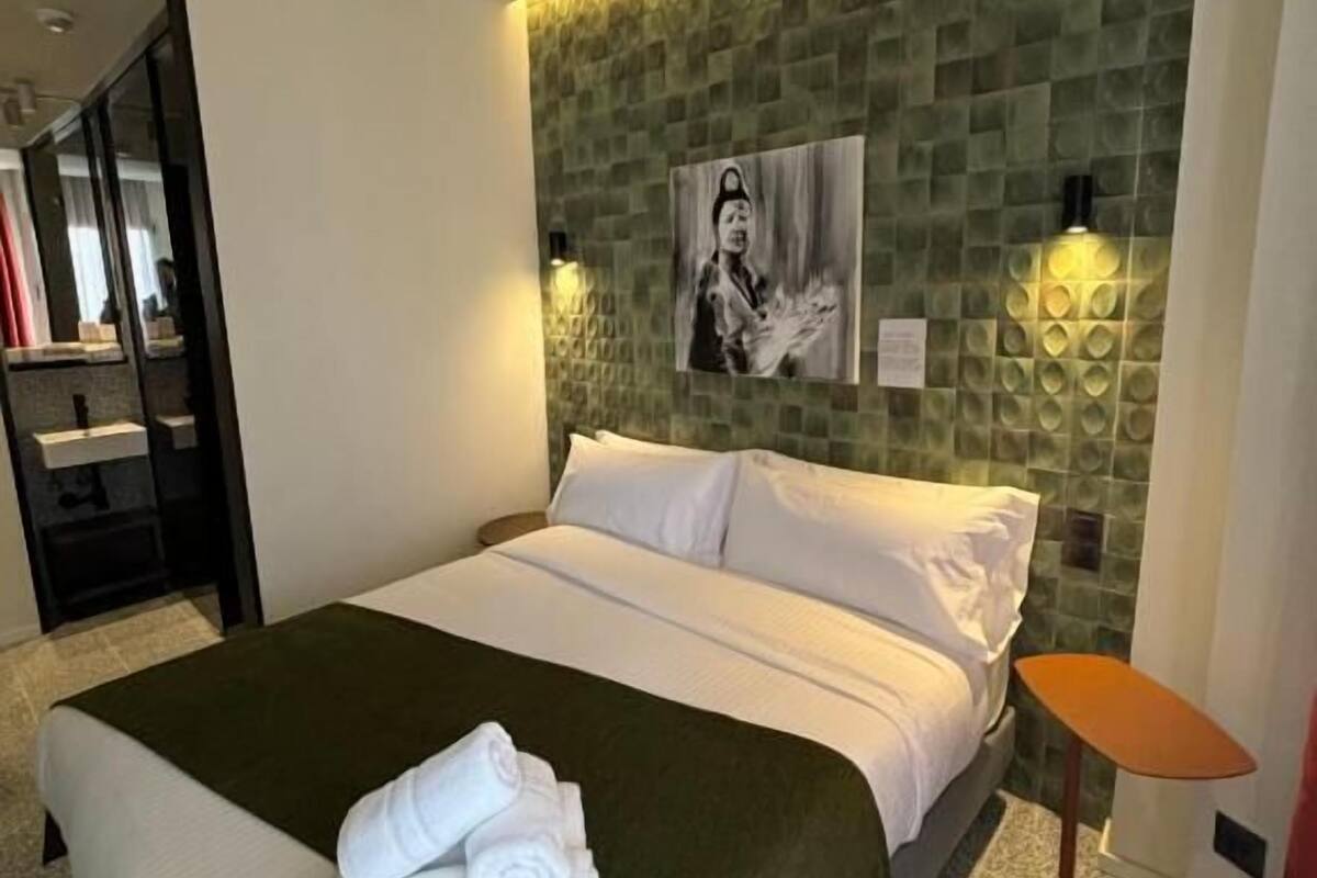 A hotel room with a bed, a green throw blanket, a folded towel, a small table, and a wall-mounted light fixture.