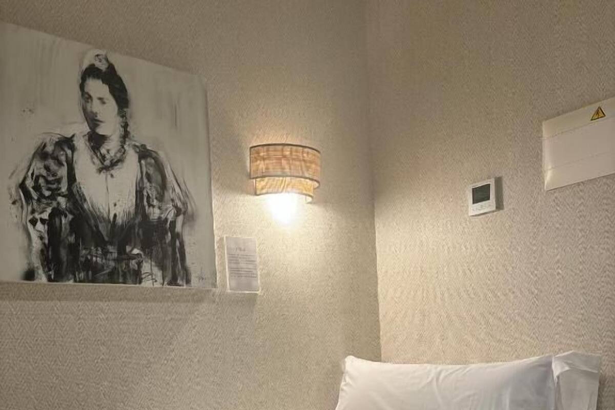 A hotel room with a bed, a green throw blanket, white pillows, a wall-mounted lamp, and a painting.