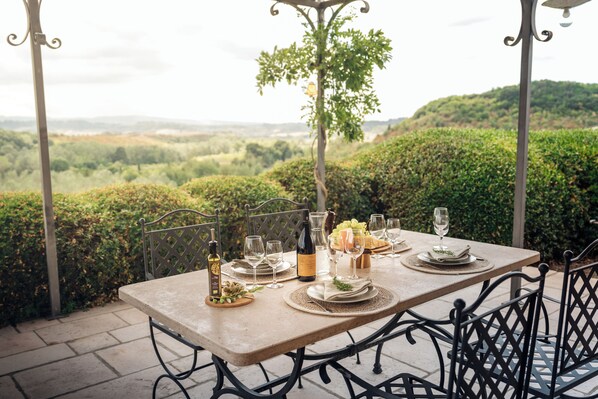 An outdoor dining table set with plates, glasses, and bottles, surrounded by metal chairs and a view of a lush landscape.