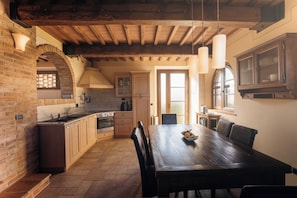 A rustic kitchen with exposed wooden beams, a brick wall, and a dining area with a wooden table and chairs.