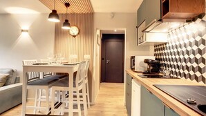 Cozinha moderna com bancada de madeira, micro-ondas embutido e máquina de café. Possui um espaço de refeições com mesa e cadeiras, e teto de madeira com luminárias pendentes.