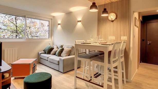Uma sala de estar moderna com área de jantar, um sofá cinza com almofadas, um banquinho redondo e um teto de madeira com luminárias pendentes.