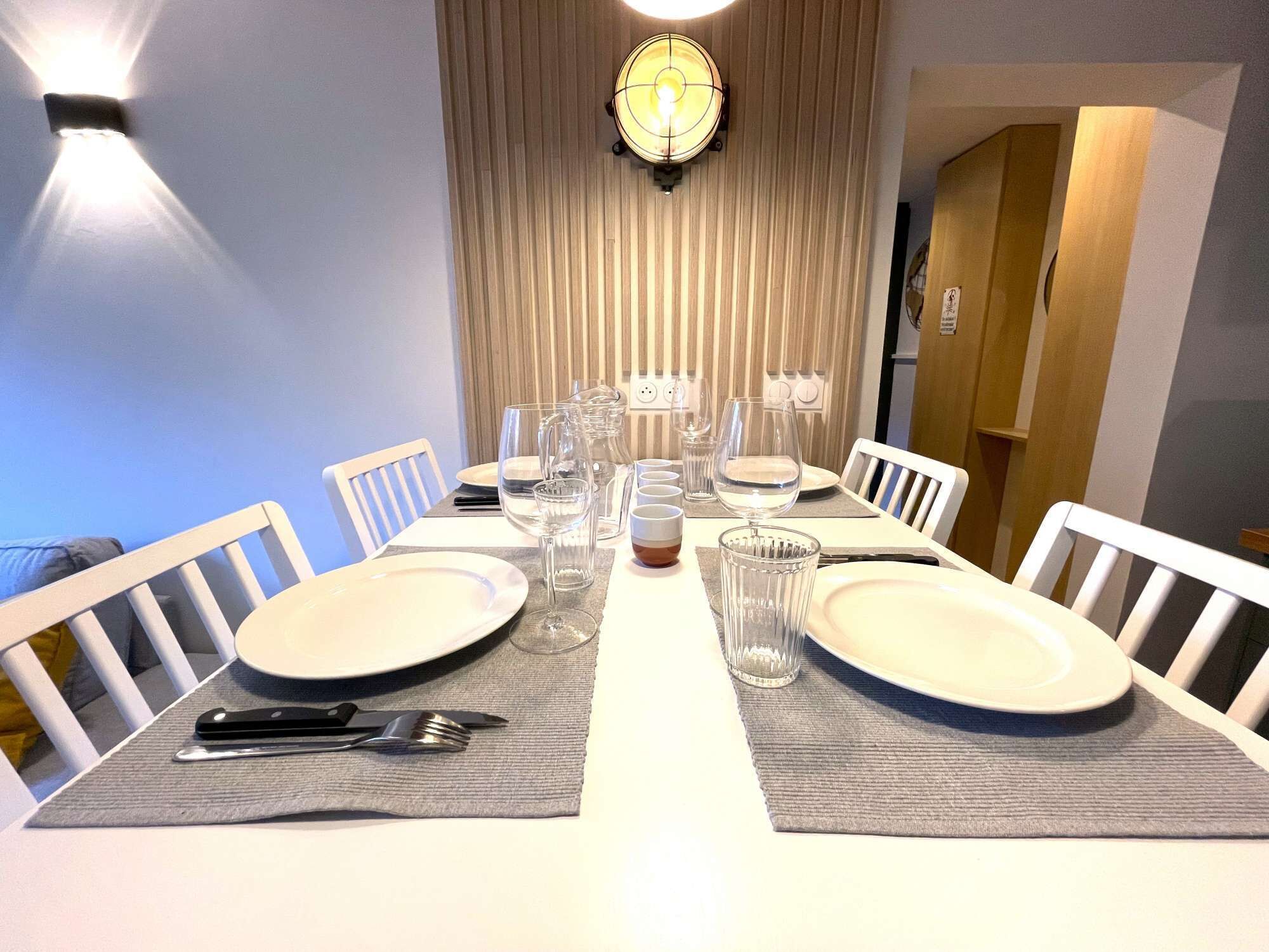 Een eettafel gedekt met borden, glazen en bestek, omringd door witte stoelen, met een wandlamp erboven.