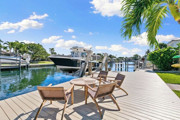 A wooden deck with outdoor furniture, a boat docked by the water, and a clear sky.