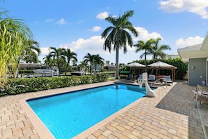 A swimming pool with lounge chairs and umbrellas, surrounded by palm trees and a brick patio.