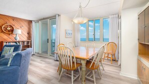 A dining area with a wooden table and chairs, a chandelier, and a view through large windows.
