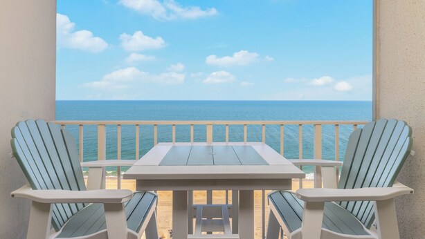 A balcony with a table and two Adirondack chairs, overlooking a beach and the ocean.