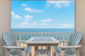Un balcon avec une table et deux fauteuils Adirondack, donnant sur une plage et l’océan.